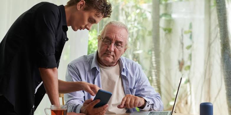 A middle-aged adult helping an elderly parent while reviewing financial documents at home.